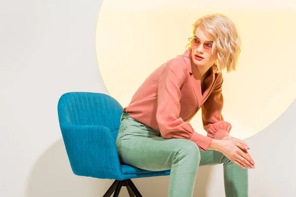 Beautiful stylish girl in colorful clothes and sunglasses sitting in armchair on white with yellow circle — Stock Photo