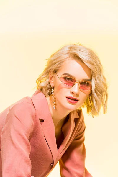 Красивая стильная блондинка в коралловой куртке смотрит в камеру и позирует на желтом — стоковое фото