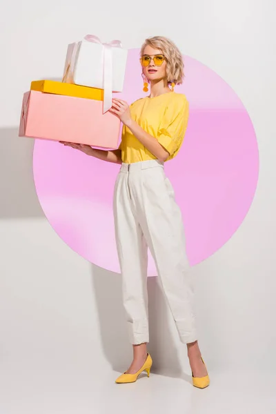 Hermosa chica elegante sosteniendo cajas de regalo y mirando a la cámara en blanco con círculo rosa - foto de stock