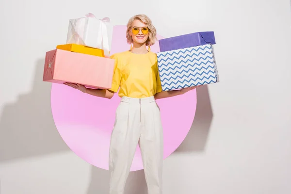 Bela menina elegante segurando caixas de presente e sorrindo no branco com círculo rosa — Stock Photo