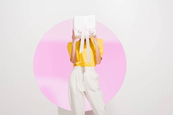Stylisches Mädchen hält Deckgesicht mit Geschenkschachtel auf weiß mit rosa Kreis — Stockfoto