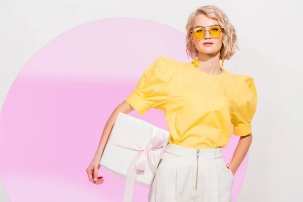 Beautiful stylish girl looking at camera and holding gift box on white with pink circle and copy space — Stock Photo