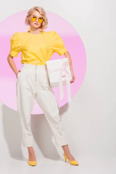 Hermosa chica con estilo mirando a la cámara y la celebración de la caja de regalo en blanco con círculo rosa — Stock Photo