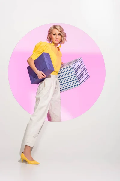 Bela menina da moda segurando caixas de presente, olhando para a câmera e posando no branco com círculo rosa — Fotografia de Stock