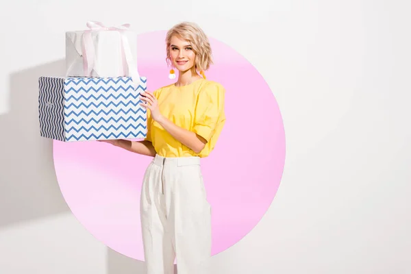 Bela menina da moda segurando caixas de presente e sorrindo no branco com círculo rosa — Fotografia de Stock