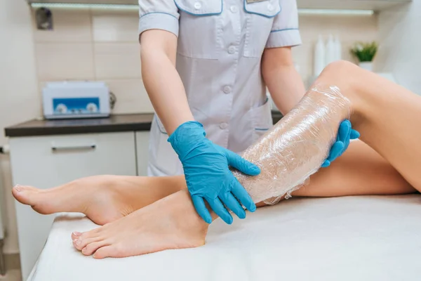 Vista recortada do cosmetologista usando filme plástico durante a sessão de embrulho corporal — Fotografia de Stock