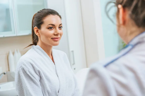 Beautiful smiling young woman in white bathrobe looking at cosmetologist in beauty clinic — Stock Photo