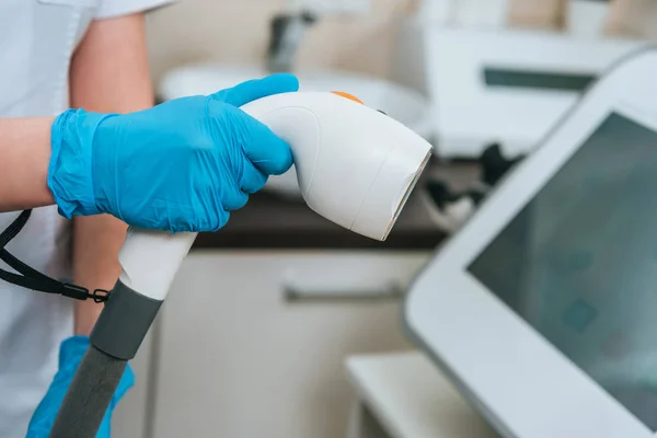 Vista recortada del cosmetólogo en guantes de goma que sostiene el dispositivo láser en la clínica - foto de stock