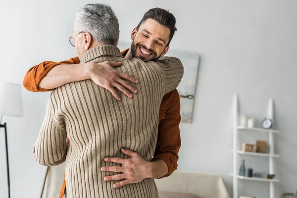 Glücklicher bärtiger Mann lächelt, während er den älteren Vater zu Hause umarmt — Stockfoto