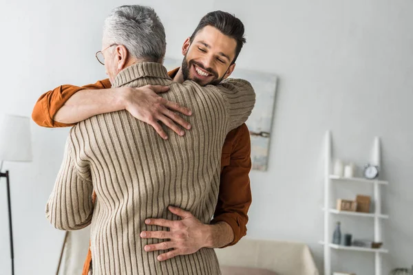 Feliz barbudo sonriendo mientras abraza padre mayor en casa - foto de stock