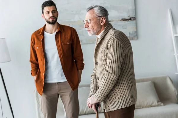 Handsome bearded son standing with hands in pockets and looking at sad senior father in glasses — Stock Photo