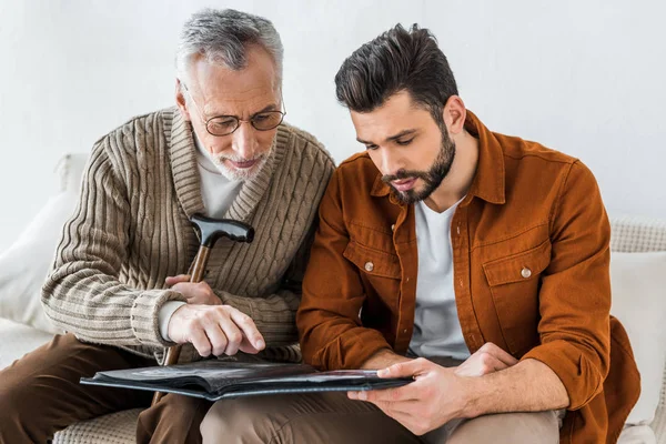 Senior father in glasses pointing with finger at photo album near son — Stock Photo
