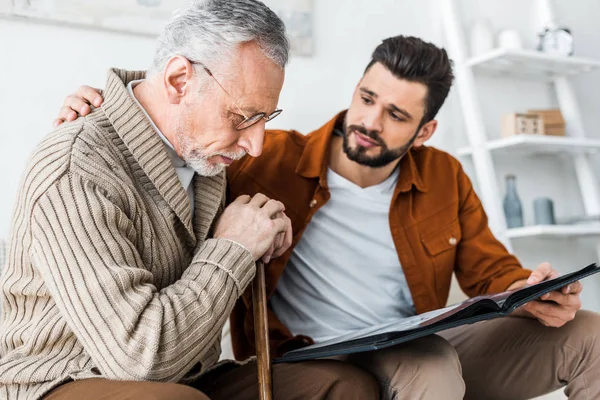 Handsome man holding photo album and looking at sad senior dad — Stock Photo