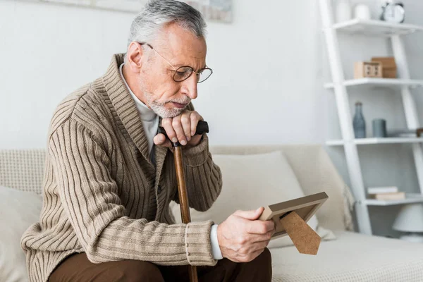 Upset retired man looking at photo frame while holding walking stick — Stock Photo