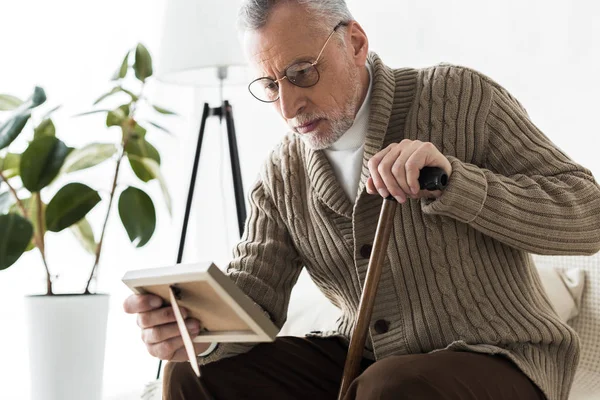 Trauriger Rentner schaut auf Fotorahmen und hält Spazierstock in der Hand — Stockfoto