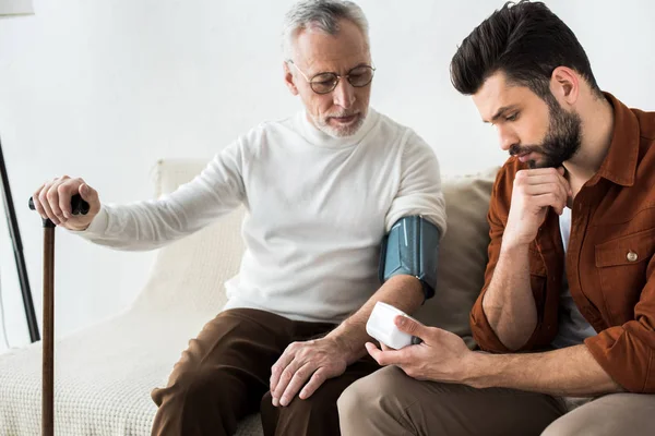 Handsome bearded man measuring blood pressure of senior father — Stock Photo