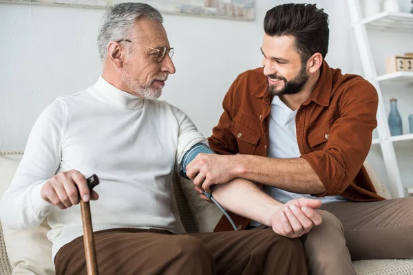 Bearded man smiling while measuring blood pressure of senior father in glasses — Stock Photo