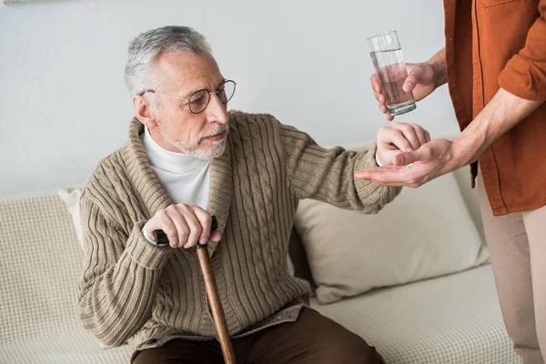 Vista recortada de hijo sosteniendo vaso de agua y pastillas cerca de padre mayor con bastón — Stock Photo