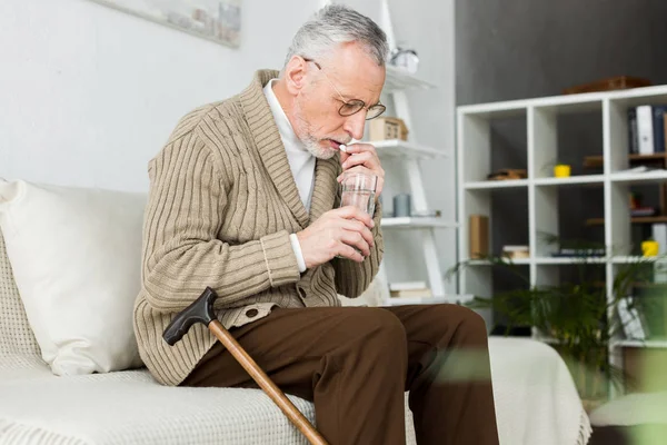 Homme âgé prenant la pilule tout en étant assis sur le canapé près de la canne à pied — Photo de stock