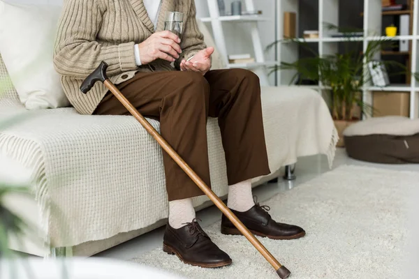 Обрезанный вид пенсионера, держащего таблетки, сидя на диване возле трости — стоковое фото