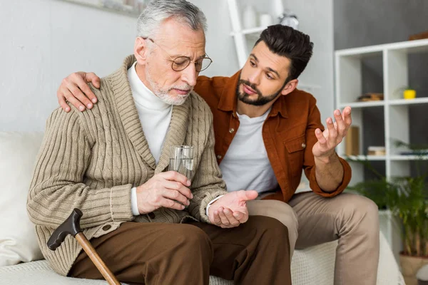 Bell'uomo gesticolare padre anziano che tiene un bicchiere d'acqua e pillola — Foto stock