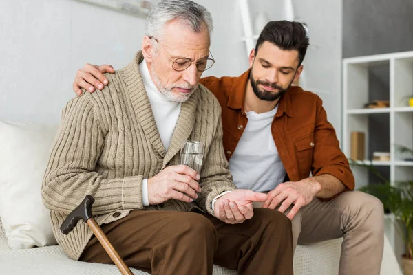 Красивий чоловік сидить з старшим батьком тримає склянку води і таблетки — стокове фото