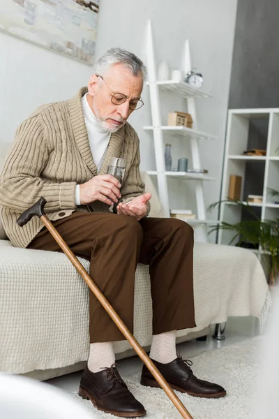 Senior man holding pill and glass of water while sitting on sofa near walking cane — Stock Photo