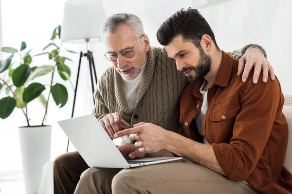 Handsome bearded man pointing with finger at laptop while sitting with senior father — Stock Photo