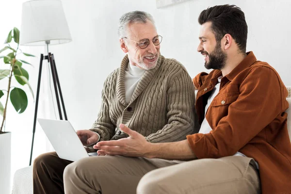 Happy bearded man gesturing while sitting with senior father and looking at laptop — Stock Photo