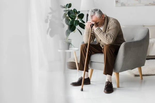 Sconvolto pensionato seduto in poltrona con bastone da passeggio a casa — Foto stock
