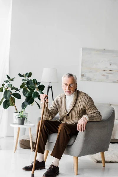 Retired man sitting in armchair with walking cane and looking at camera — Stock Photo