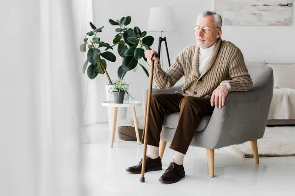 Пенсионер в очках сидит в кресле с тростью — стоковое фото