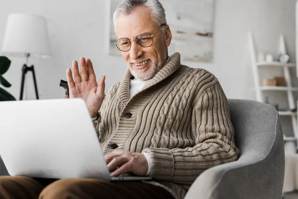 Cheerful senior man in glasses waving hand while having video call — Stock Photo