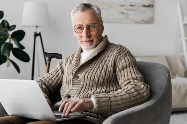 Senior mit Brille blickt in Kamera, während er mit Laptop zu Hause sitzt — Stockfoto