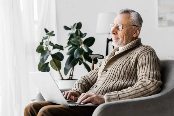 Heureux homme âgé dans des lunettes en utilisant un ordinateur portable à la maison — Photo de stock