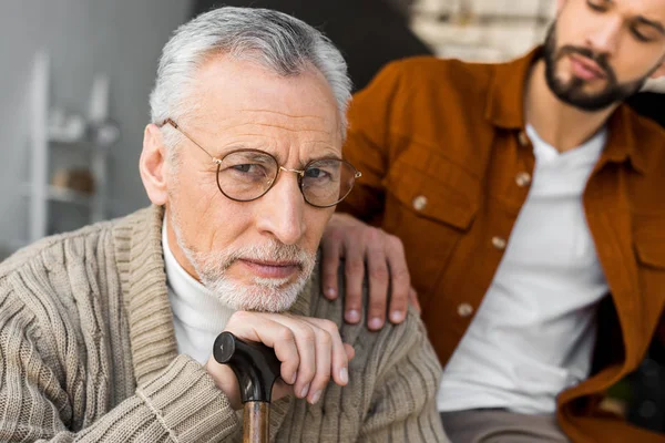 Vista ritagliata del figlio barbuto mettere mano sulla spalla del padre anziano triste in occhiali — Foto stock