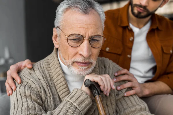 Ausgeschnittener Blick auf Sohn, der aufgebrachten Senior-Vater die Hände in Brille auf die Schultern legt — Stockfoto
