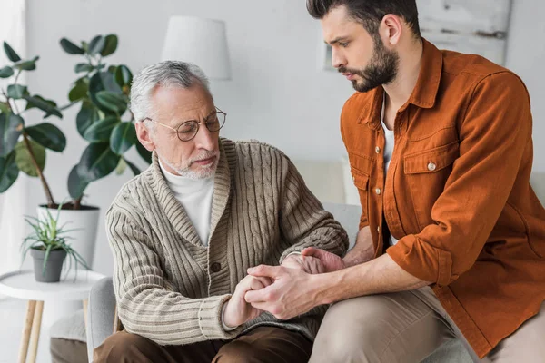 Padre mayor molesto en gafas cogidas de la mano con el hijo guapo en casa - foto de stock