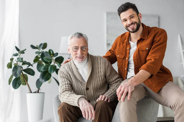 Glücklicher älterer Vater mit Brille sitzt neben gutaussehendem und fröhlichem Sohn zu Hause — Stockfoto