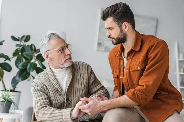 Sad senior father in glasses holding hands with handsome son at home — Stock Photo