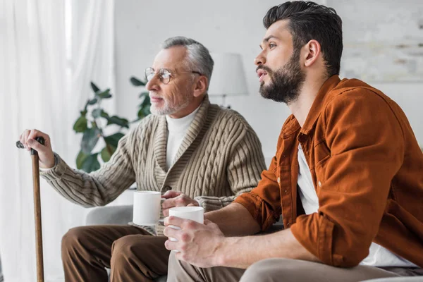 Pensionierter Vater in Brille sitzt mit gutaussehendem Mann und hält Tasse — Stockfoto