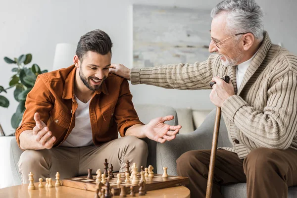 Père à la retraite dans des lunettes mettant la main sur l'épaule de fils heureux tout en jouant aux échecs — Photo de stock