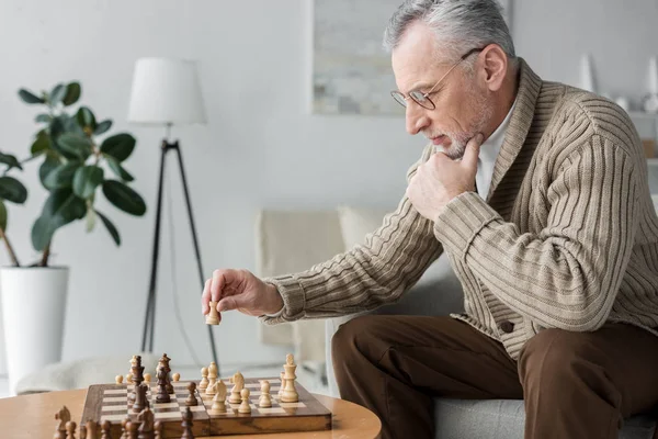 Retired man in glasses thinking while playing chess at home — Stock Photo