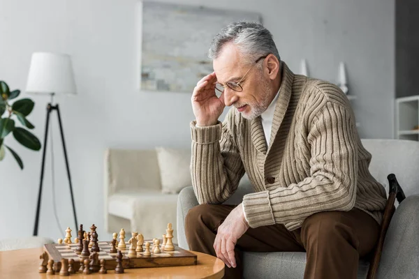 Pensive retired man in glasses thinking while playing chess at home — Stock Photo