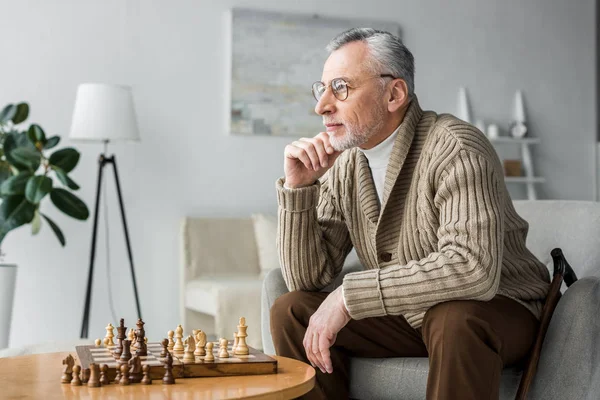 Thoughtful retired man in glasses thinking while sitting near chess board at home — Stock Photo