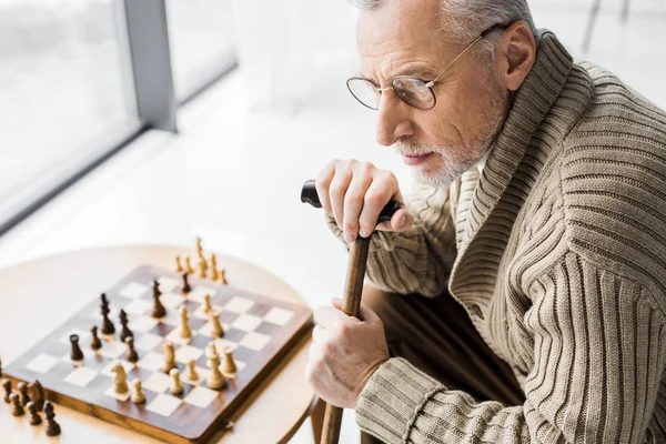 Vista aerea di pensionato in occhiali pensando mentre seduto vicino a scacchiera a casa — Foto stock