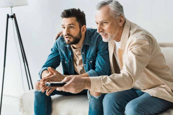 Alegre padre mayor sosteniendo mando a distancia cerca apuesto hijo mientras mira la televisión en casa — Stock Photo