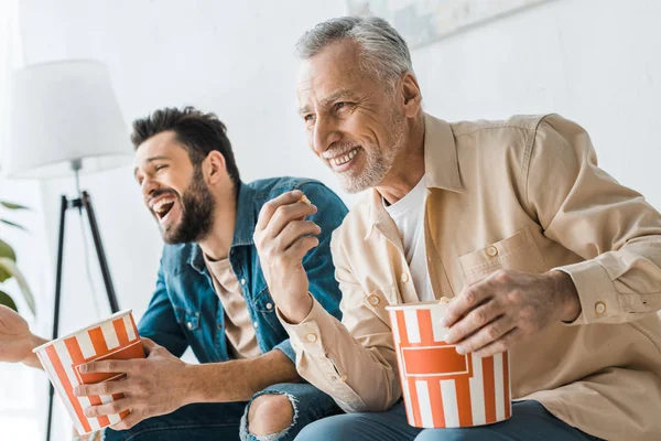 Happy senior father sitting with cheerful son and holding popcorn bucket — Stock Photo