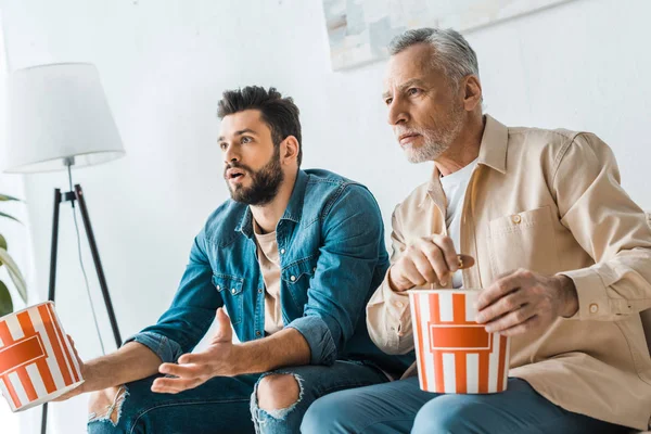 Padre mayor sorprendido sentado con hijo guapo y sosteniendo palomitas de maíz cubo mientras mira la televisión - foto de stock