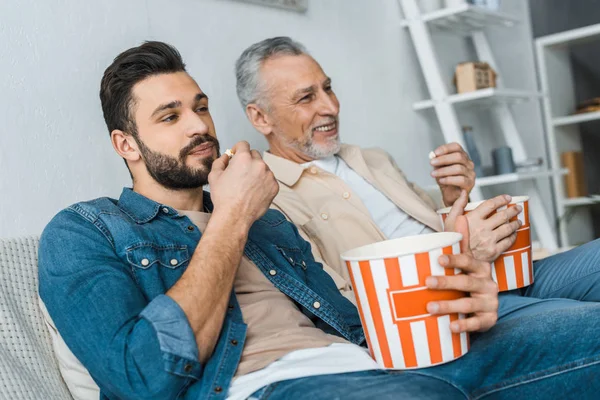 Feliz padre mayor sentado con hijo guapo y sosteniendo palomitas de maíz cubo mientras mira la televisión - foto de stock